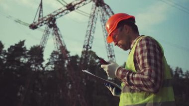 Elektrik mühendisi, başlık ve güvenlik yeleği giyiyor, yüksek voltajlı elektrik hatlarında elektrik sistemlerini kontrol ediyor, telsizle konuşuyor, müfettişin çalışma planı panosu var. Elektrik şirketi çalışanı
