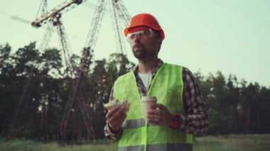 Hungry worker in overalls in electric power business eats sandwich and drinks takeaway coffee at workplace near power lines. Power worker eating snack with hot drink near high voltage power line