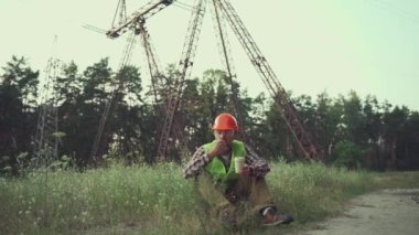 Engineer having lunch near power lines and electricity pylons in the field. An electrical substation worker in a protective helmet eats a sandwich and drinks coffee during a break at the workplace