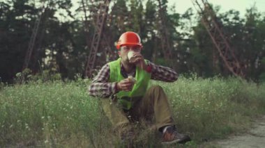 Lunch time. Worker in hard hat holding coffee cup and sandwich during lunch break background of power lines in field. Worker on work site having meal. Electrical engineer eating snack with drink