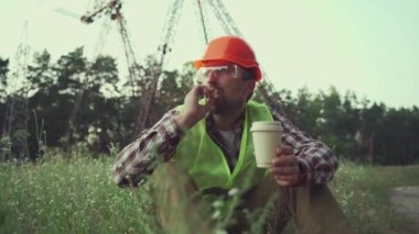 Hungry worker in overalls in electric power business eats sandwich and drinks takeaway coffee at workplace near power lines. Power worker eating snack with hot drink near high voltage power line