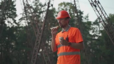Engineer having lunch near power lines and electricity pylons in the field. An electrical substation worker in a protective helmet eats a sandwich and drinks coffee during a break at the workplace