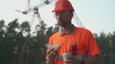 Lunch time. Worker in hard hat holding coffee cup and sandwich during lunch break background of power lines in field. Worker on work site having meal. Electrical engineer eating snack with drink