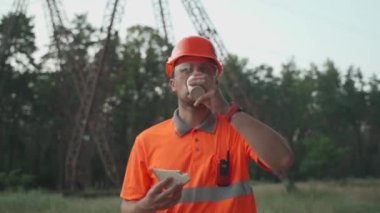 Hungry worker in overalls in electric power business eats sandwich and drinks takeaway coffee at workplace near power lines. Power worker eating snack with hot drink near high voltage power line
