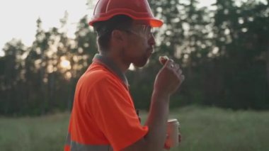 Engineer having lunch near power lines and electricity pylons in the field. An electrical substation worker in a protective helmet eats a sandwich and drinks coffee during a break at the workplace