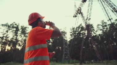 Öğle yemeği molasında inşaat işçisi. Teknisyen, kasklı bir mühendis öğle yemeği yiyor ve işyerindeki elektrik hattı altyapısıyla kahve içiyor. Elektrikçi abur cubur yer ve kahve içer.