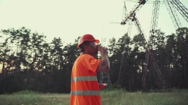 Hungry worker in overalls in electric power business eats sandwich and drinks takeaway coffee at workplace near power lines. Power worker eating snack with hot drink near high voltage power line — Stockvideo