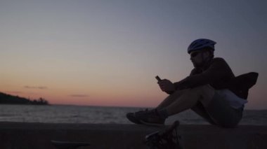 Sağlıklı, aktif bir yaşam tarzı ve bisiklet yolculuğu. Kafkasyalı adam denize bisikletle gitti ve akıllı telefon navigatörüyle rota çizmek için durdu. Deniz kenarında gün batımında bisikletçi rüzgarlı havada akıllı bir telefon kullanır.