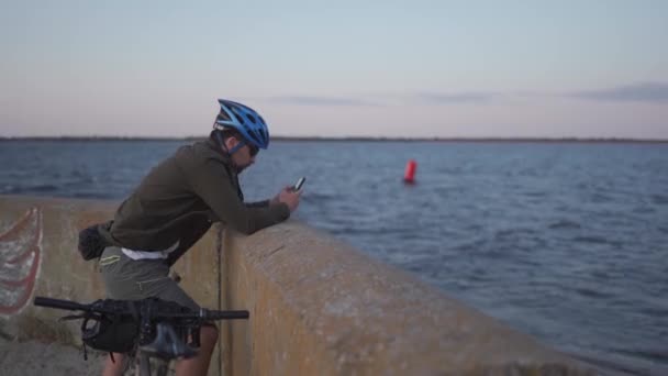 Tema è il ciclismo in riva al mare. Ciclista di sesso maschile si è fermato per godersi la vista sul mare e vedere la strada sulla mappa sul suo smartphone. Un maschio con una bicicletta sul terrapieno sta riposando e utilizzando uno smartphone — Video Stock