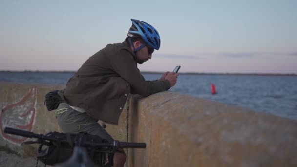 健康的で活発なライフスタイルとサイクリング旅行。白人男性は自転車に乗って海に行き、スマートフォンのナビゲーターでルートをプロットするのを止めた。夕暮れ時の海のサイクリストは、風の強い日にスマートフォンを使用します — ストック動画