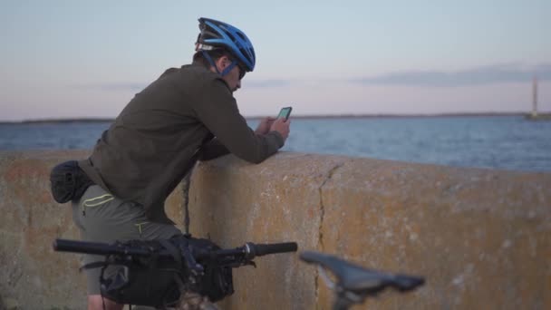 Cyklistický výlet na moře při západu slunce. Muž cyklista odpočívá na nábřeží po moři a používá smartphone vybudovat trasu a navigaci GPS. Muž na kole na mořském pobřeží surfování na internetu na chytrý telefon — Stock video