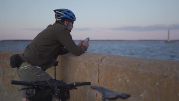 男性サイクリストは海沿いの遊歩道で休憩し、日没時にスマートフォンを使用します。自転車で健康的なアクティブなライフスタイル旅行者。自転車道のある海岸線に立つ自転車道、地図を見る電話 — ストック動画