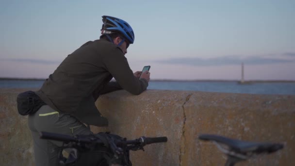 Thema is fietsen in zee. Man fietser gestopt om te genieten van het uitzicht op zee en zie de weg op de kaart op zijn smartphone. Een man met een fiets op de dijk rust uit en gebruikt een smartphone — Stockvideo