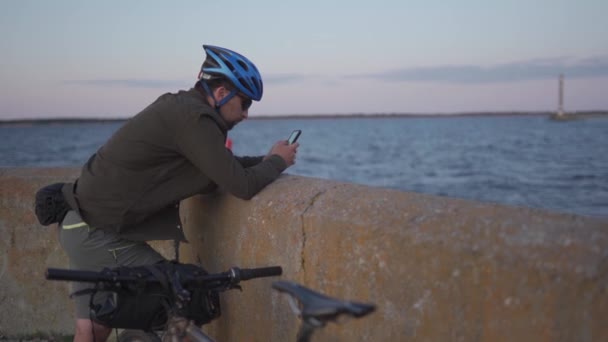 Estilo de vida saudável e ativo e viagens de bicicleta. Homem caucasiano andava de bicicleta para o mar e parou para traçar rota no navegador do smartphone. Ciclista ao pôr do sol pelo mar usa um telefone inteligente em tempo ventoso — Vídeo de Stock