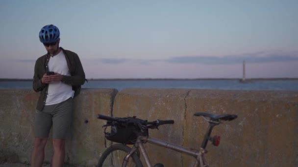Mannelijke fietser rust op promenade over zee en maakt gebruik van smartphone bij zonsondergang. Gezonde actieve lifestyle reiziger op de fiets. Fietsenmaker staat aan de kust zeezicht met fiets te bouwen route, kijkt naar kaart op de telefoon — Stockvideo