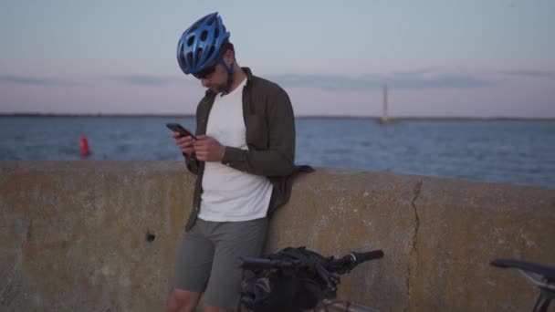 Tema es el ciclismo en la playa. Hombre ciclista se detuvo para disfrutar de la vista al mar y ver la carretera en el mapa en su teléfono inteligente. Un hombre con una bicicleta en el terraplén está descansando y usando un teléfono inteligente — Vídeo de stock