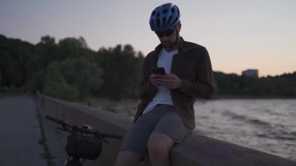 Temaet er sykling i havet. En mannlig syklist stoppet for å nyte sjøutsikten og se veien på kartet på smarttelefonen sin. En mann med sykkel på fyllingen hviler og bruker smarttelefon. – stockvideo
