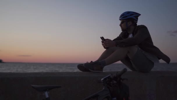 Viaje en bicicleta al mar al atardecer. Hombre ciclista está descansando en terraplén por mar y utiliza el teléfono inteligente para construir la ruta y navegar GPS. Hombre con bicicleta en la orilla del mar navegar por Internet en un teléfono inteligente — Vídeos de Stock