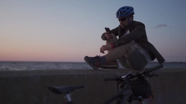 Ciclista di sesso maschile che riposa sul lungomare via mare e utilizza smartphone al tramonto. Un sano viaggiatore dello stile di vita attivo in bicicletta. Bicicletta si trova sulla costa vista mare con percorso di costruzione di biciclette, guarda la mappa sul telefono — Video Stock