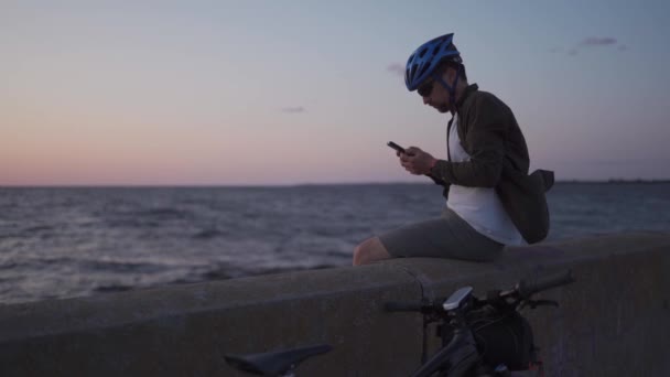 Thema ist das Radfahren am Meer. Der männliche Radfahrer hielt an, um den Meerblick zu genießen und die Straße auf der Landkarte auf seinem Smartphone zu sehen. Ein Mann mit Fahrrad am Bahndamm ruht sich aus und benutzt ein Smartphone — Stockvideo