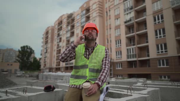 Voorman, arbeider of architect van een mannelijke bouwer die op de bouwplaats thee of koffie drinkt in een afhaalbekertje en met een mobiele telefoon praat. Rust maakt deel uit van het productiviteitspakket — Stockvideo