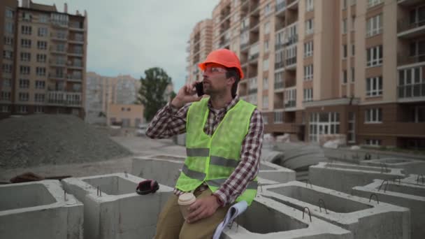 Man byggmästare talar i telefon under lunchrasten. Byggarbetare ringer på smarttelefon under kaffepausen på byggarbetsplatsen. Konstruktören dricker kaffe och ger instruktioner till underordnade — Stockvideo