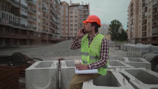 No hay tiempo para descansar. Constructor en casco y chaleco en café funciona hablando por teléfono con los contratistas. Ingeniero de construcción está bebiendo café para llevar y haciendo una llamada en su teléfono celular — Vídeo de stock