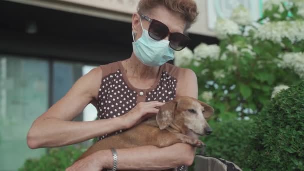 Mujer anciana en máscara médica se sienta en el banco de la ciudad y desinfecta a su perro manos salchichas y patas con antiséptico. Mujer mayor enmascarada con un desinfectante para mascotas en el parque durante la cuarentena covid 19 — Vídeos de Stock