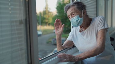 90 'ların sonlarında maskeli yaşlı bir kadının portresi ev penceresinden el sallıyor. Koruyucu maskeli yaşlı kadın pencereden dışarı bakar ve evde yalnızken elini sallar ve sosyal mesafeyi yönetir..