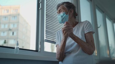 Üst düzey kadın el dezenfektanı sürüyor, Covid-19 'a karşı koruyucu maske takıyor, pencerenin önünde duruyor. Evde el antibakteriyel sıvı dezenfektanı kullanan, yüzü maskeli, üzgün, yaşlı bir kadın..