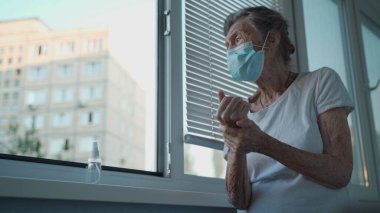 Tıbbi maskeli yaşlı bir kadın alkol jeliyle ya da antibakteriyel sabun dezenfektanıyla bir huzurevinin penceresinde ellerini yıkıyor. COVID-19 koronavirüs salgını sırasında hijyen.