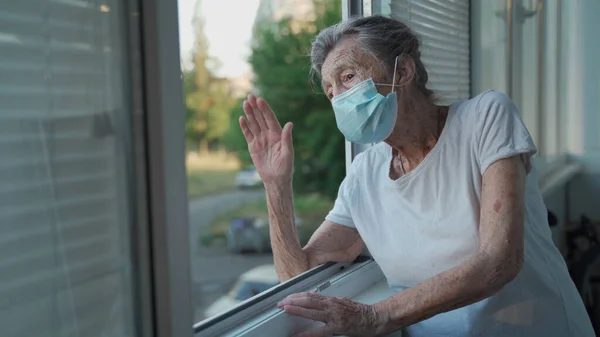 Portrait of masked senior woman in late 90s waving from home window. Elderly woman in protective mask looks out the window and waves her hand while isolating at home and conducting social distancing.