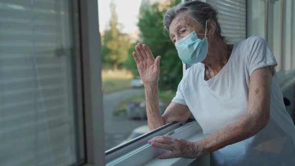 Happy senior woman in mask waves to family from window hospital. Masked elderly female glad that children came visit her in nursing home. Grandmother greets child with hand of relatives at clinic.