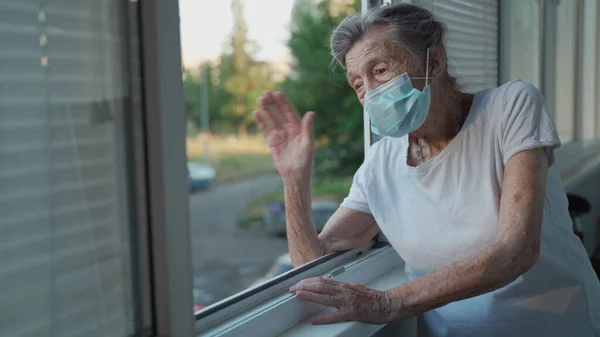 Happy senior woman in mask waves to family from window hospital. Masked elderly female glad that children came visit her in nursing home. Grandmother greets child with hand of relatives at clinic.