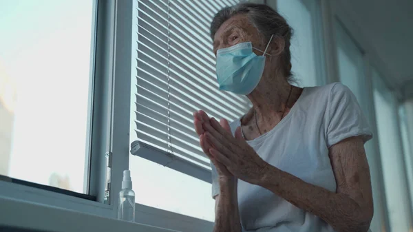 Elderly woman in a medical mask washing hands with alcohol gel or antibacterial soap sanitizer standing by the window in a nursing home. Hygiene during the COVID-19 coronavirus pandemic.