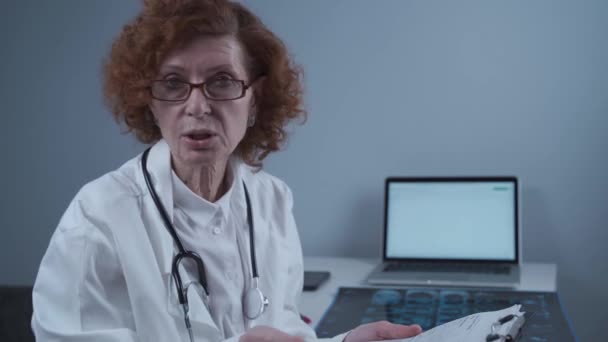 Professional elderly woman doctor holding clipboard, talking about diagnosis while looking at camera in hospital office. Consulting and therapy concept. Healthcare, senior clinic staff, medical help — Stock video