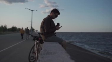 Traveler on bicycle stopped to enjoy the view and check road map on smartphone to the sea and have coffee in cycle time of travel. Male cyclist use smartphone and drinks takeaway coffee on embankment