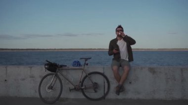 Active rest and healthy lifestyle theme. The cyclist stopped to rest, drink coffee and surf the Internet on the phone while sitting on the promenade by the sea during a bike ride. Coffee break