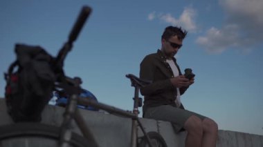 Man cyclist resting overlooking sea, drinking coffee and using smartphone while sitting on embankment. Male 30s wearing sunglasses, drinking takeaway coffee and using mobile phone on waterfront