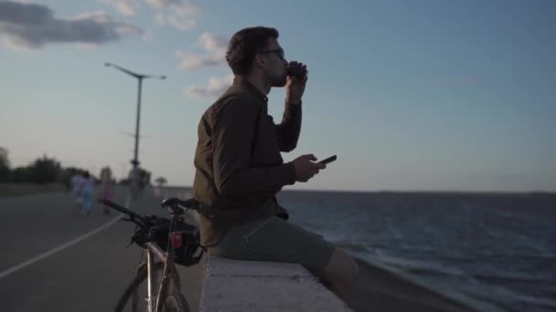 Man cyclist resting overlooking sea, drinking coffee and using smartphone while sitting on embankment. Male 30s wearing sunglasses, drinking takeaway coffee and using mobile phone on waterfront — Video Stock
