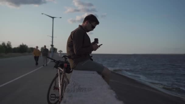 El viajero en bicicleta se detuvo para disfrutar de la vista y comprobar la hoja de ruta en el teléfono inteligente hacia el mar y tomar un café en el tiempo de ciclo de viaje. Hombre ciclista uso de teléfono inteligente y bebidas para llevar café en terraplén — Vídeo de stock