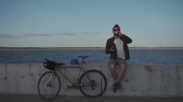 Active rest and healthy lifestyle theme. The cyclist stopped to rest, drink coffee and surf the Internet on the phone while sitting on the promenade by the sea during a bike ride. Coffee break — Stok video