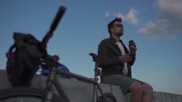 Traveler on bicycle stopped to enjoy the view and check road map on smartphone to the sea and have coffee in cycle time of travel. Male cyclist use smartphone and drinks takeaway coffee on embankment — Αρχείο Βίντεο