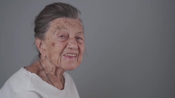 Senior kvinna 90 år gammal ler, skrattar och visar en protes på en grå bakgrund. Äldre kvinna med djupa rynkor och grått hår glad i en bra humör studio porträtt tittar på kameran — Stockvideo