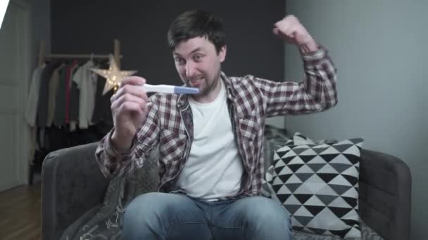 Future young father is very happy about news of pregnancy, holds pregnancy test and is incredibly happy at home on the couch. Happy excited guy looking at positive pregnancy test and celebrating — Stock Video