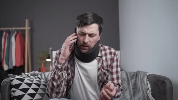 Man in foam cervical collar talking on phone, upset with news. Male in cervical brace calls at insurance company for medical assistance. Person after car accident with spinal injury suffering at home — Stock Video