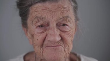 90 yaşında, beyaz dişsiz yaşlı bir kadının portresi. Derin kırışıklıkları ve gri saçları var. Stüdyoda gülümsüyor ve gri arka planda konuşuyor. Diş sorunları olan yaşlı bir kadın. Ortodontist hasta