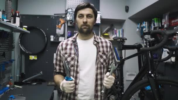 Bicicleta loja mecânico engraçado posando com ferramentas em mãos olhando para a câmera. Trabalhador da loja de bicicletas está orgulhosamente na frente do inventário com instrumentos e se divertindo posando em sua loja de reparação de bicicletas — Vídeo de Stock