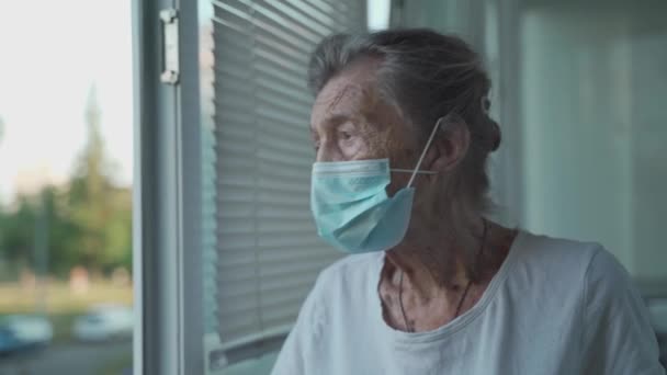 一位悲伤的老年妇女，90岁，戴着防护口罩，凝视着疗养院开着的窗户。脸上戴着面具的孤独老年女性焦虑地站在窗边，举手面对 — 图库视频影像