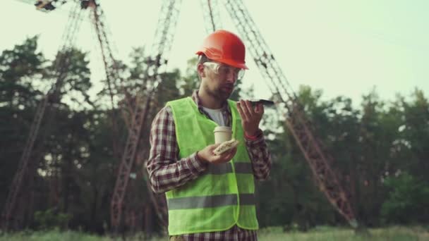 Engenheiro elétrico usando chapéu duro grita com seu telefone em desagrado durante o almoço no local de trabalho da subestação. Linha de transmissão elétrica trabalhador bebe café e come sanduíche durante videochamada — Vídeo de Stock